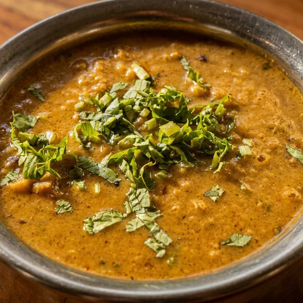 A close-up of a metal bowl filled with a rich, golden-brown lentil stew, garnished with fresh green herbs.