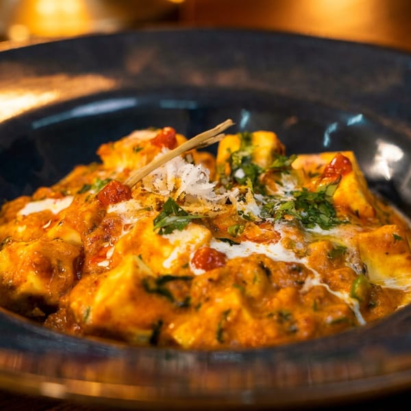 A vibrant orange curry dish with paneer, garnished with fresh herbs and shredded coconut, in a dark bowl.