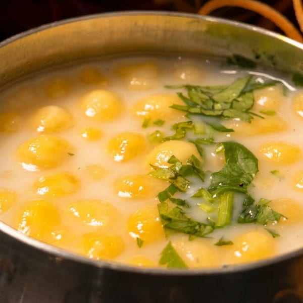 A metal bowl filled with yellow chickpea dumplings in a white sauce, garnished with green herbs.