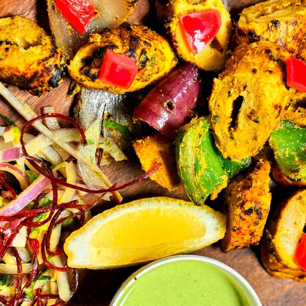 Grilled chicken tikka with bell peppers, onions, lemon, and green chutney on a wooden plate.