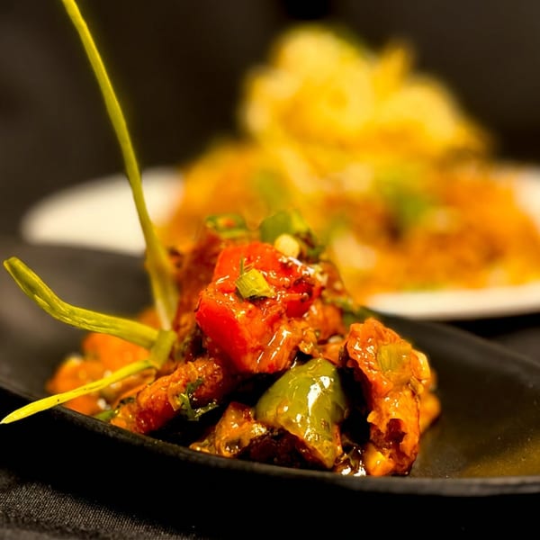 Spicy chicken stir-fry with bell peppers and scallions on a black plate, with rice in the background.